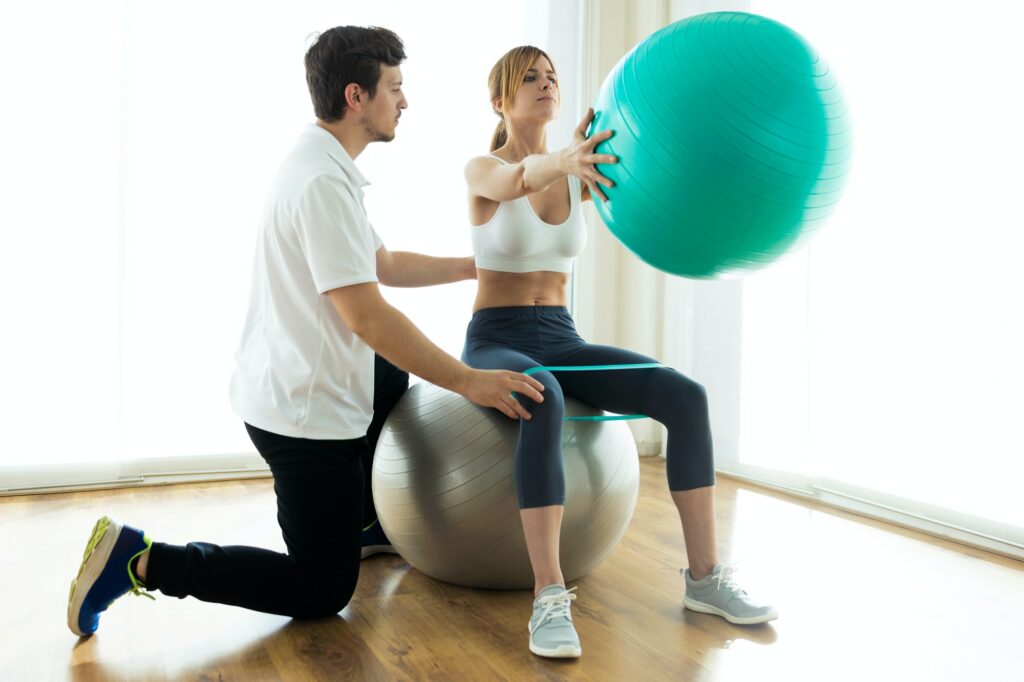 Physiotherapist helping patient to do exercise on fitness ball in physio room.